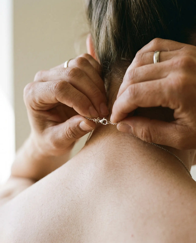 Two hands fastening a fine chain clasp at the back of the neck
