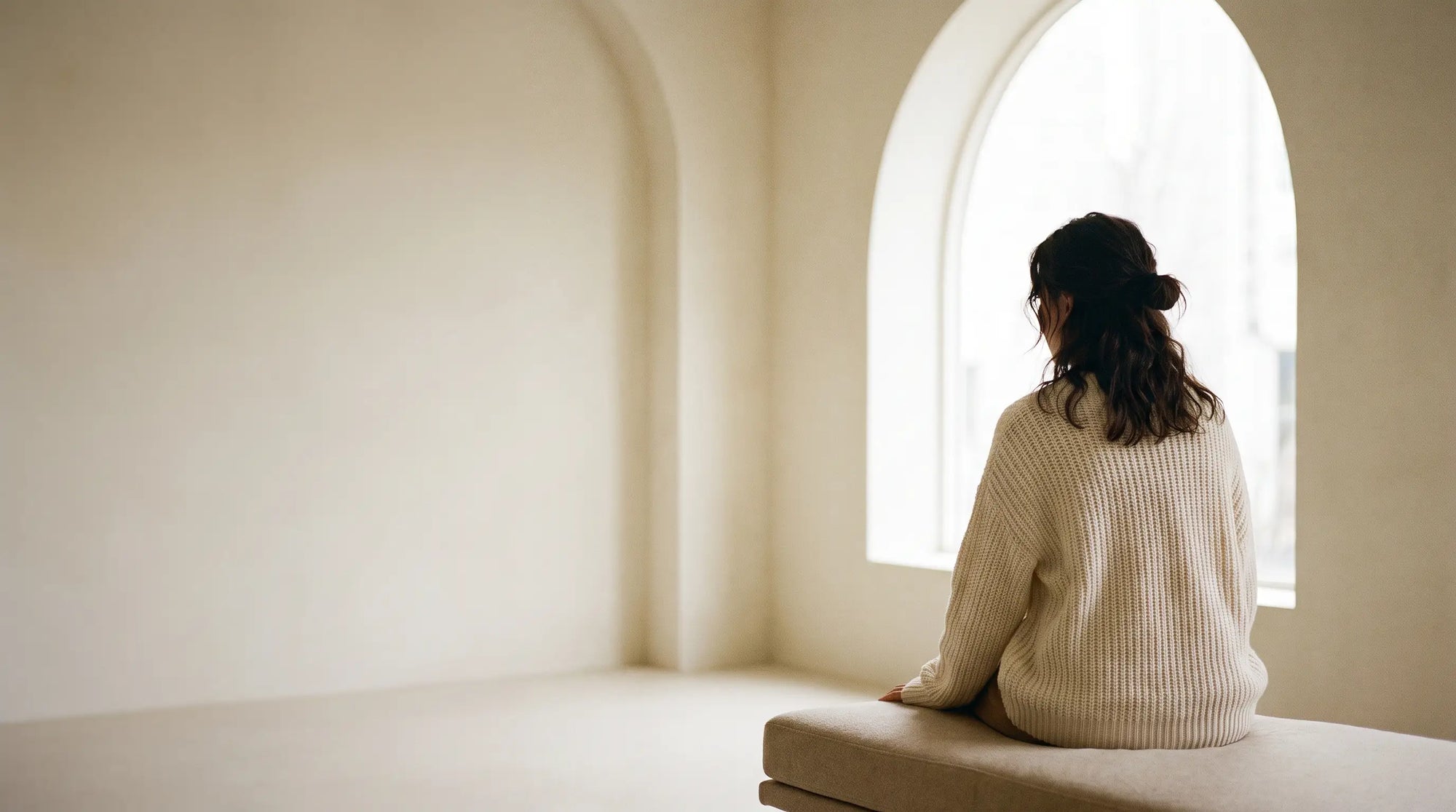 A woman seated by a bright arched window in soft cream knitwear